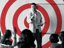 Graphic showing a black and white photo of a teacher at the front of a classroom filled with students with a red bullseye target over the teacher