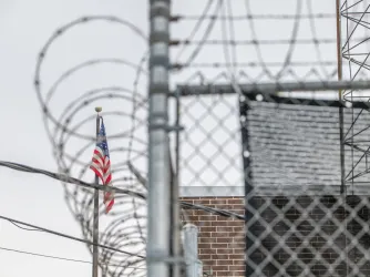 Photo of the ICE processing facility in Broadview, Illinois, showing a chainlink fence topped with razorwire surrounding the building and an American flag in the background.