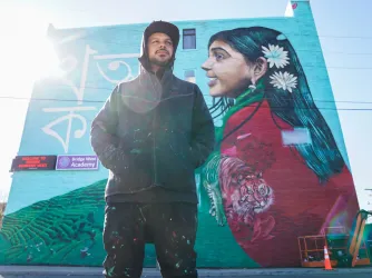 Artist Victor MARKA27 Quinonez poses before his mural on Bangladeshi life on the side of Bridge Academy West in Detroit Michigan. (Image via Imagn / USA Today images)