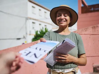 Woman wearing a hat and green shirt hands out flyers in a sunny urban setting