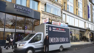 Emerson College student Sam Neves standing in front of a billboard truck with a FIRE ad that says "Emerson Kinda Sus"