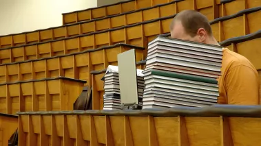 Student in lecture hall with face hidden behind books and laptop