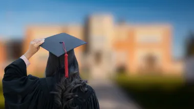 A graduate in cap and gown stands with their back to the camera facing a blurred campus building
