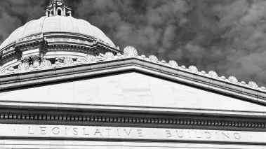 Black and white exterior of Legislative chambers of the Washington State Capitol in Olympia with inscription, pillars, and dome with a cloudy sky in the background.