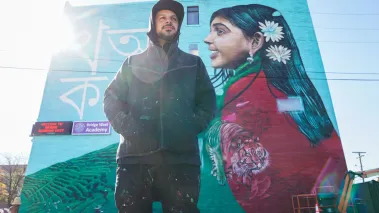 Artist Victor MARKA27 Quinonez poses before his mural on Bangladeshi life on the side of Bridge Academy West in Detroit Michigan. (Image via Imagn / USA Today images)
