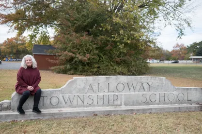 FIRE client Gail Nazarene sitting on a stone sign that reads "Alloway Township Schools"