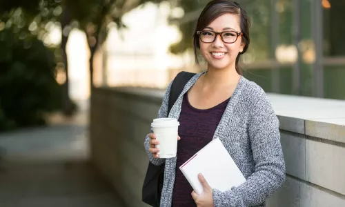 university student smiling with coffee and book bag on campus