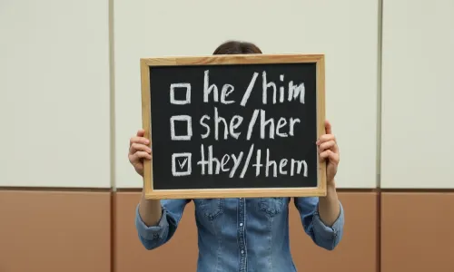 Woman holding chalkboard with list of gender pronouns near wall.jpg