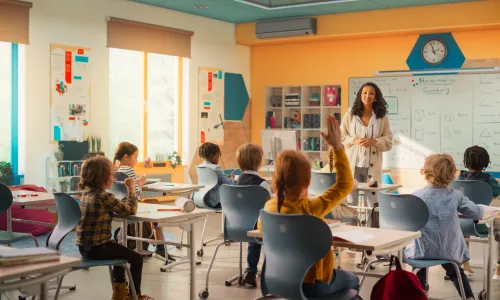 Teacher explaining a math lesson to classroom full of children and one girl is raising her hand