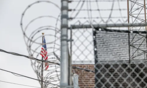 Photo of the ICE processing facility in Broadview, Illinois, showing a chainlink fence topped with razorwire surrounding the building and an American flag in the background.