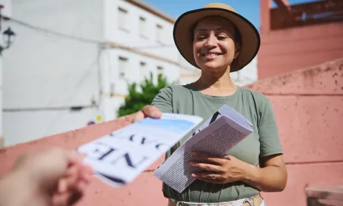 Woman wearing a hat and green shirt hands out flyers in a sunny urban setting