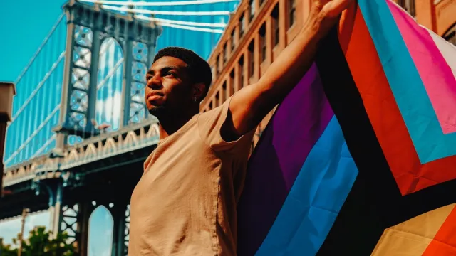 Black man holds Progress Pride flag in front of the Manhattan Bridge